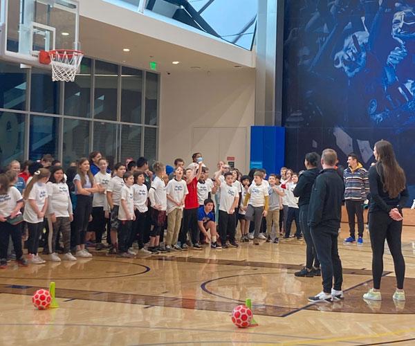 Arco Iris 4th and 5th grade students listen to Nike employees as they prepare to play a game. They are inside a gymnasium at the Nike World Headquarters.