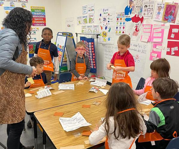 Kindergarten students at Arco Iris Immersion School participate in a Home Depot building event in their classroom. The students are wearing orange Home Depot aprons while sitting at tables and working with wood kits. An adult is offering help.