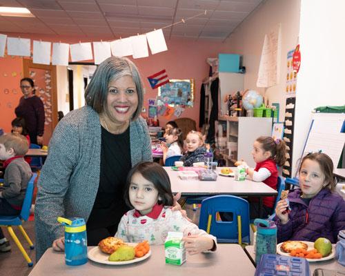 Arco Iris Spanish Immersion School teacher Angie Tirado smiles for a photo with 2 kindergarten students eating lunch in the classroom.